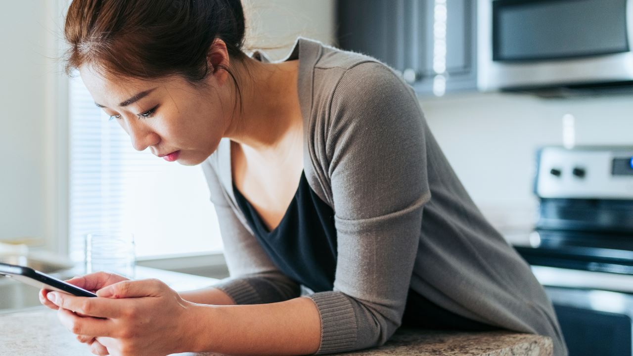 Asian woman leaning on her kitchen counter while looking at her smartphone